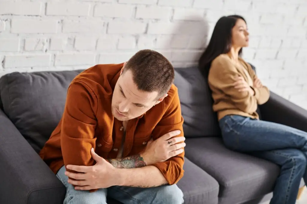 gloomy tattooed man sitting on couch in living room near offended asian wife, family problem